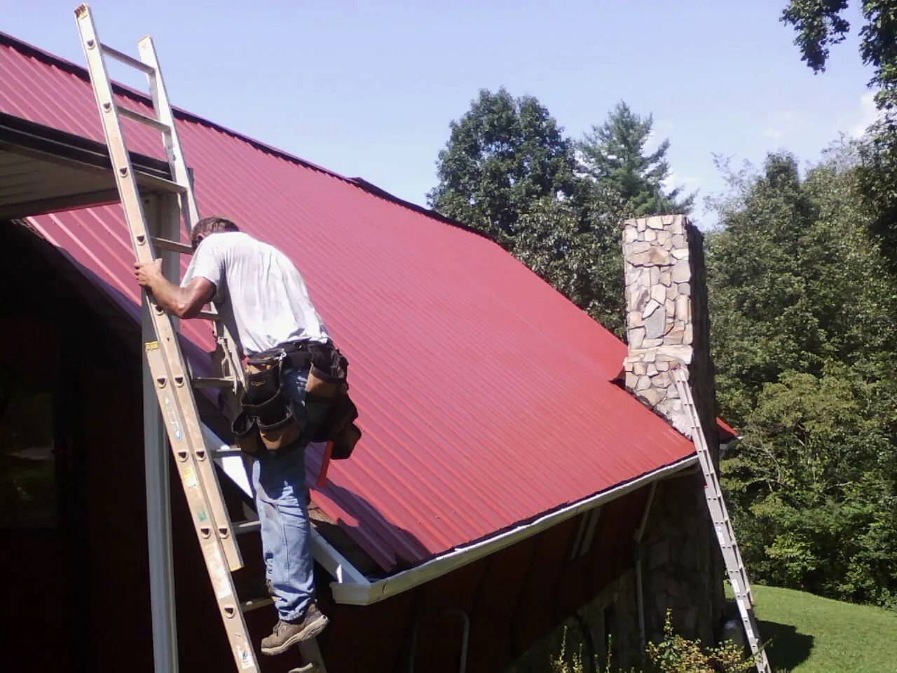 Roofer on a ladder beside a red metal roof and stone chimney — Soffit & Fascia Repair by Roofing Brothers in Letts